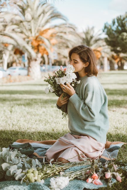 A young woman sitting on a patterned blanket in a park with tall palm trees and a clear sky in the background. She is holding a fresh floral bouquet consisting of white and soft pink roses, white asti