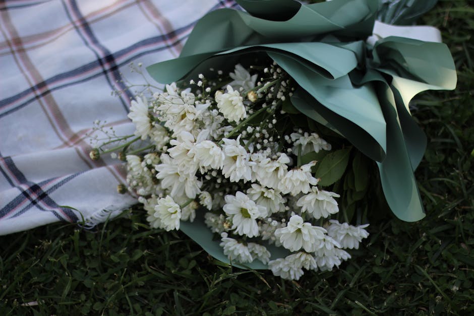 A bouquet of fresh white chrysanthemums and small white filler flowers, wrapped in matte green paper with a white ribbon, rests on a grassy surface beside a plaid cloth. The flowers display layered pe