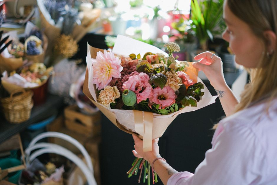 A woman with long blond hair, wearing a light pink shirt, is holding a bouquet of fresh flowers in a flower shop. The bouquet includes large pink dahlias, soft pink lisianthus, creamy carnations, and 