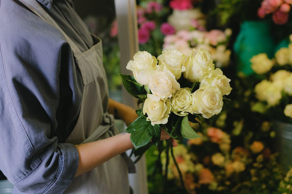 A person dressed in a grey floral apron holds a bouquet of fresh, creamy white roses with dark green leaves. The roses are tightly clustered, with smooth petals and sturdy stems, wrapped in tissue pap