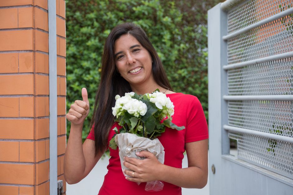 A young woman with long dark hair, smiling warmly and giving a thumbs-up gesture, is holding a potted plant with white flowers wrapped in brown paper. She is wearing a bright red short-sleeved top and