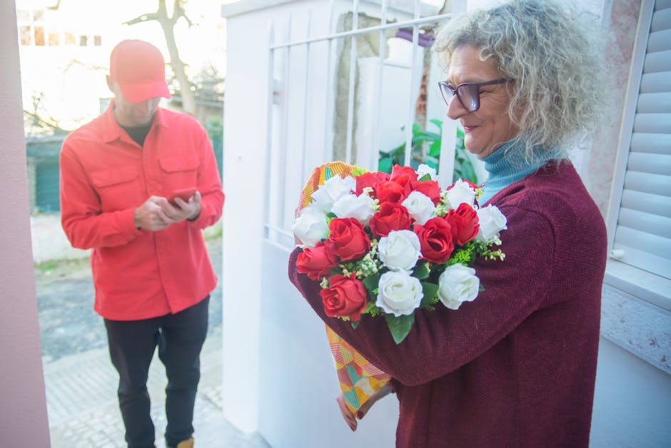 A woman with curly gray hair, wearing glasses, a blue turtleneck, and a red sweater, is holding a vibrant bouquet of fresh red and white roses with green foliage, wrapped in colorful patterned paper.